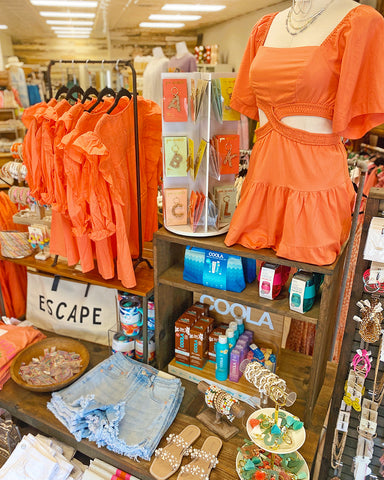 Rockwall Store front table display featuring denim shorts, an orange cutout dress, an orange shirt, accessories and other travel necessities